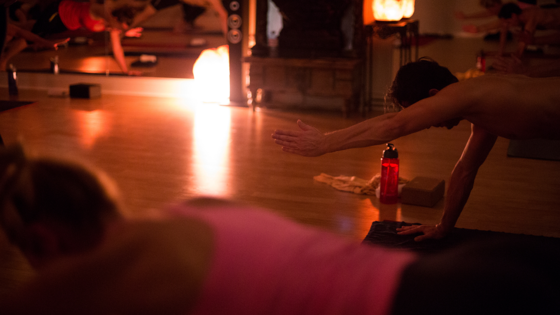 Dim, warm-lit yoga studio with students stretching on mats; a red water bottle and towel sit beside a mat in the foreground.
